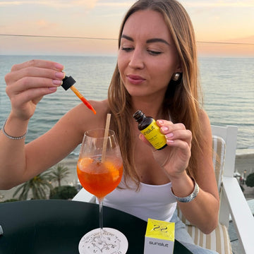 Woman adding Sunslut Glowjob No.1 tanning drops to an Aperol spritz during sunset by the beach.